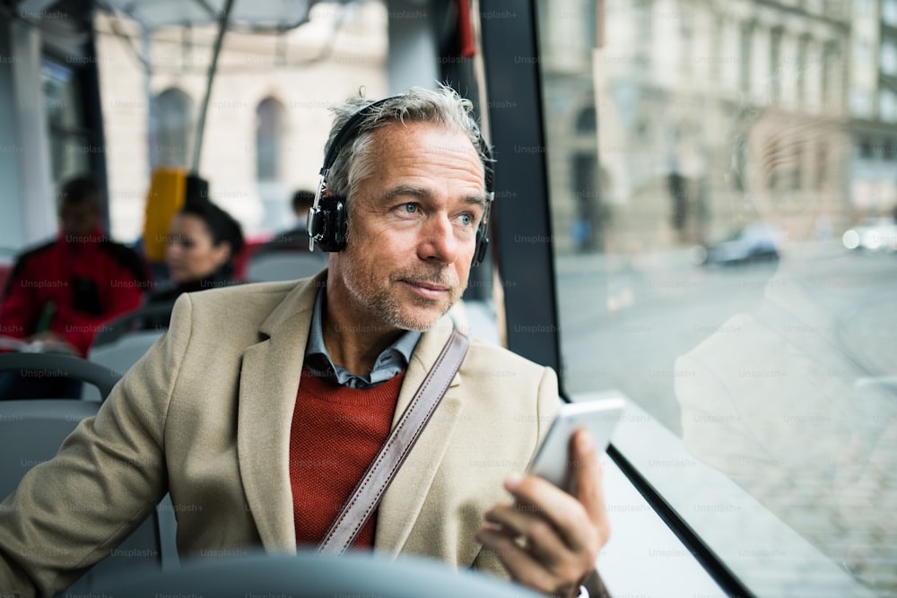 Empresário maduro e cansado com smartphone e heaphones viajando de ônibus na cidade, ouvindo música.