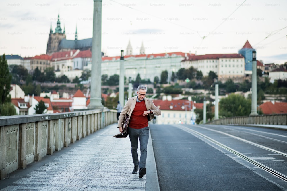 Mature handsome businessman with bag walking on a bridge in Prague city, checking the time. Copy space.