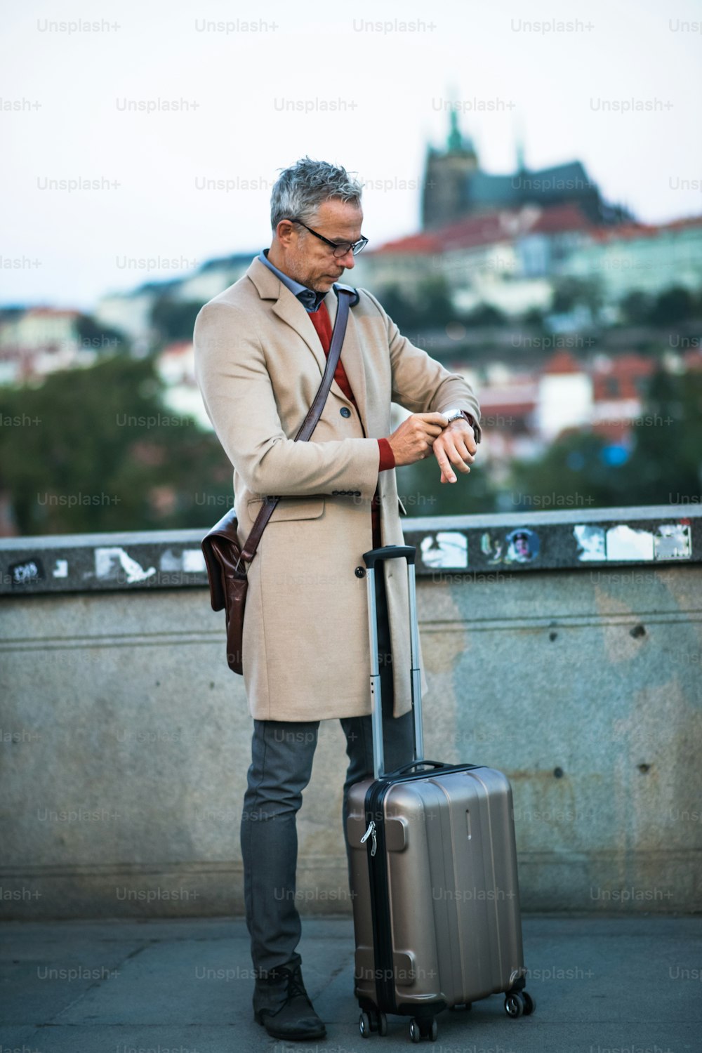 Reifer, gutaussehender Geschäftsmann mit Koffer, der auf einer Brücke in Prag steht und die Zeit überprüft. Speicherplatz kopieren.