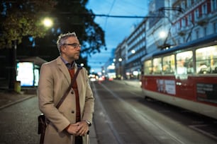 Mature businessman with suitcase waiting for a tram or a trolley car in the evening in Prague city.