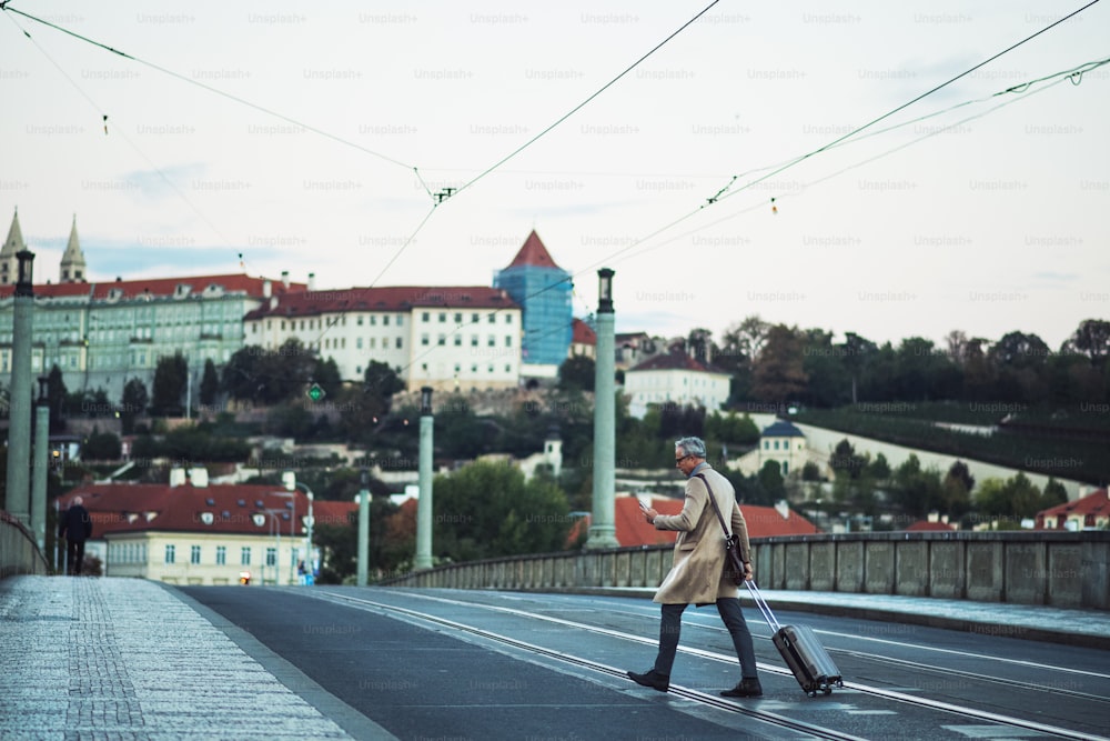スマートフォンとスーツケースを持つ成熟したビジネスマンがプラハ市の道路を横断し、テキストメッセージ。