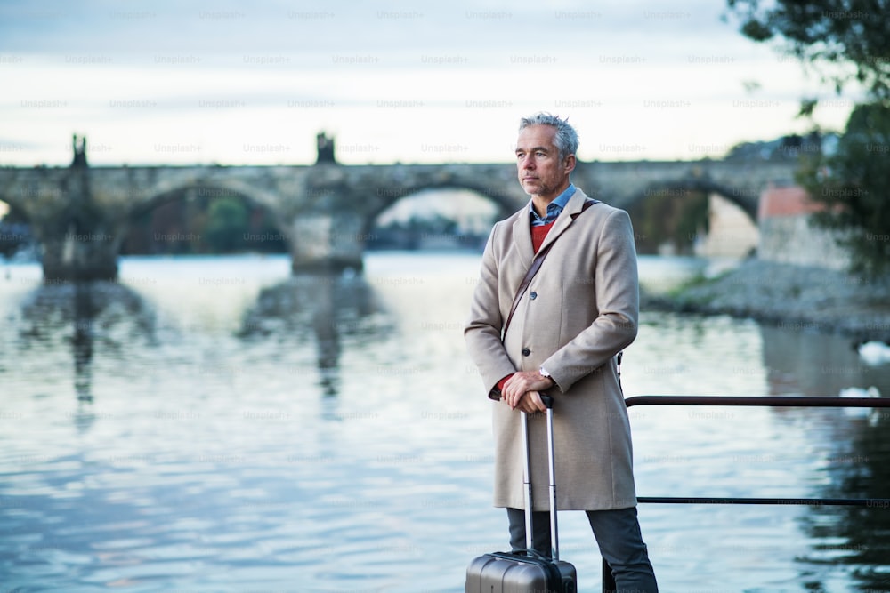 Empresário bonito maduro com mala de pé junto ao rio Vltava na cidade de Praga. Espaço de cópia.