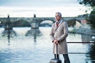 Maturo bell'uomo d'affari con valigia in piedi vicino al fiume Moldava nella città di Praga. Copia spazio.