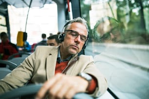 Mature tired businessman with glasses and heaphones travellling by bus in city, listening to music.
