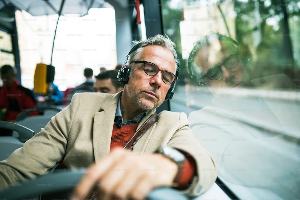 Mature tired businessman with glasses and heaphones travellling by bus in city, listening to music.