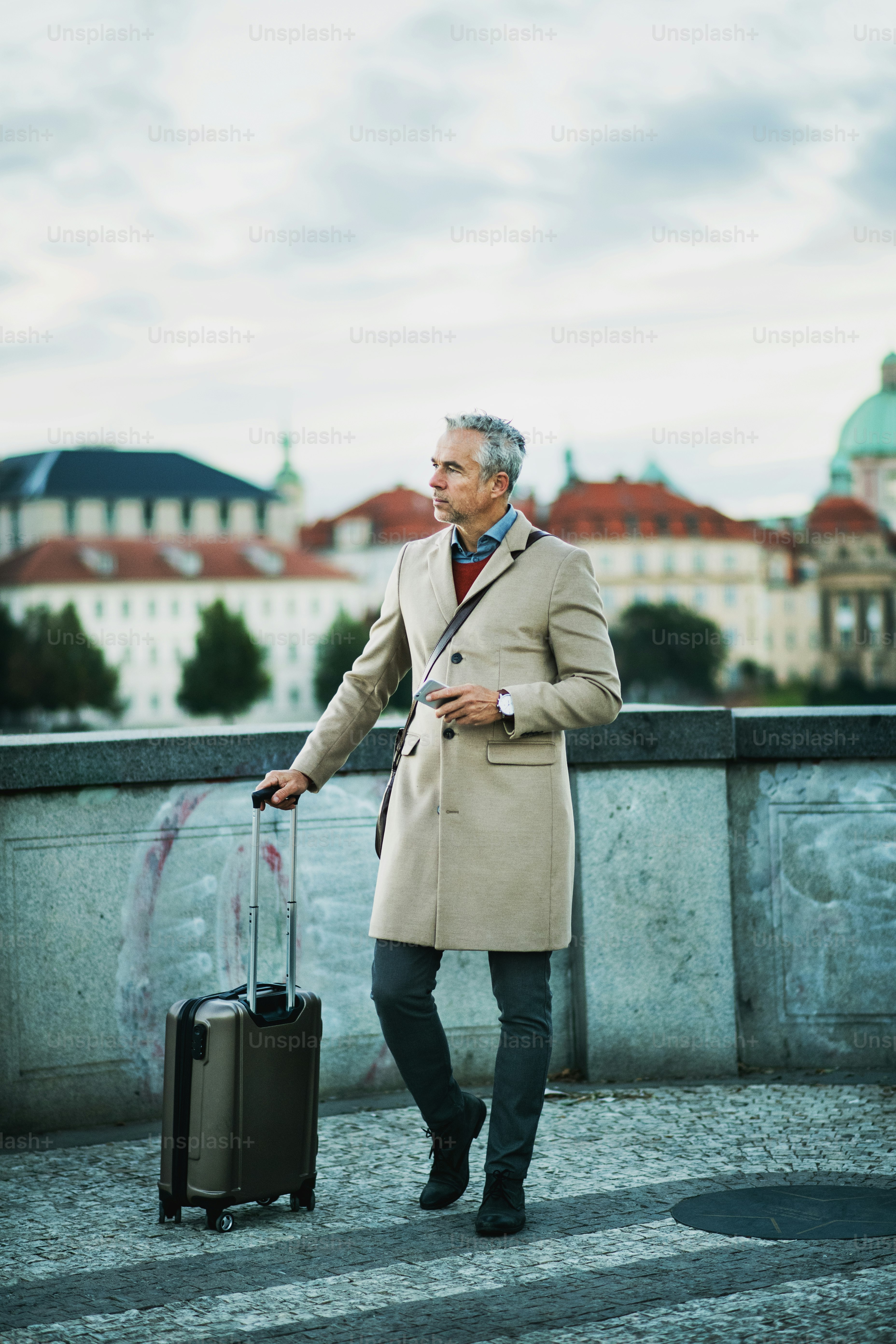 Mature handsome businessman with suitcase and smartphone standing on a bridge in Prague city. Copy space.