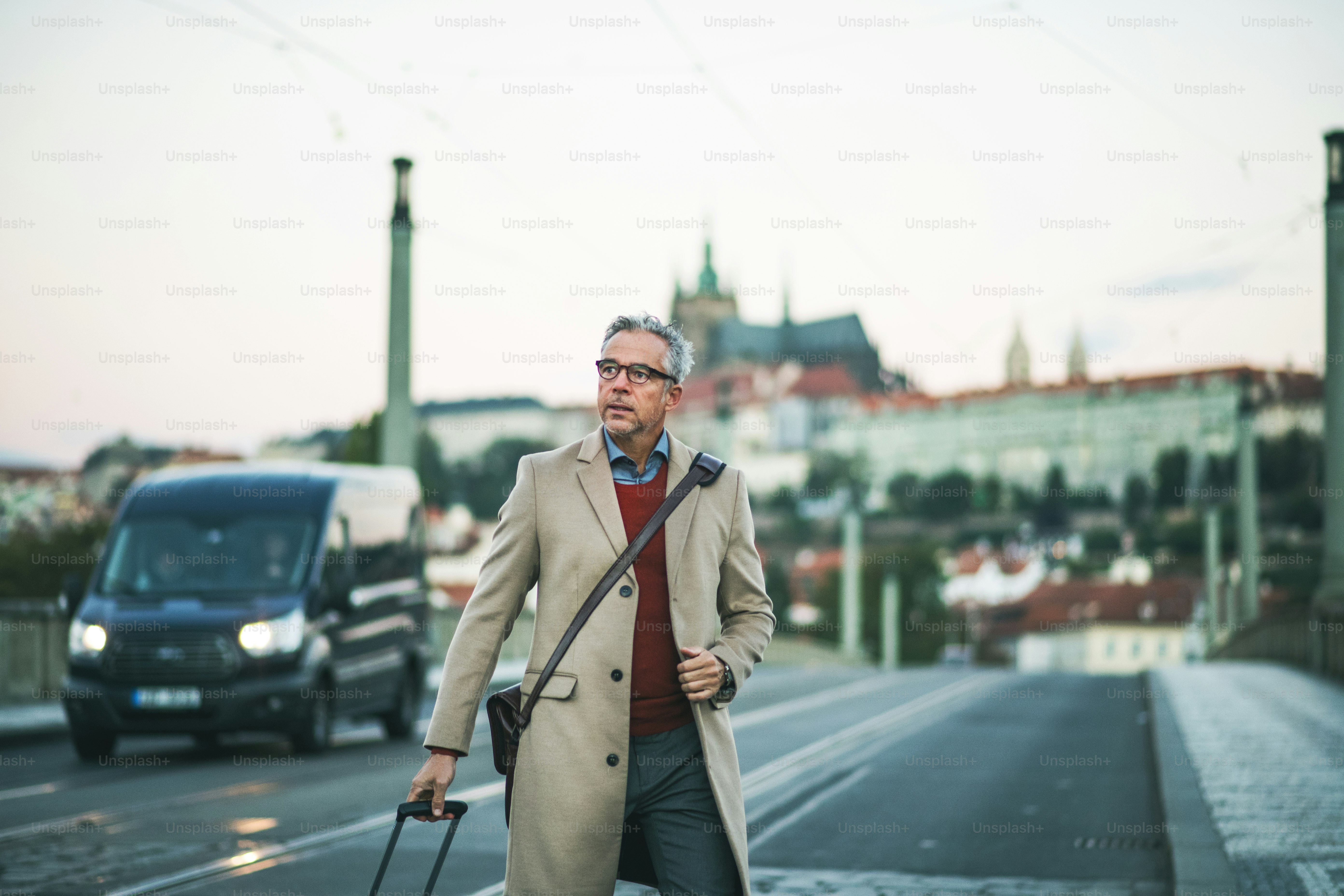 Mature handsome businessman with suitcase walking on a bridge in Prague city, crossing the road. Copy space.