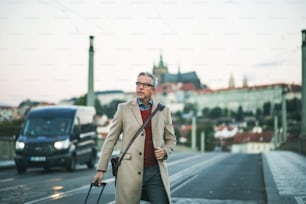 Mature handsome businessman with suitcase walking on a bridge in Prague city, crossing the road. Copy space.