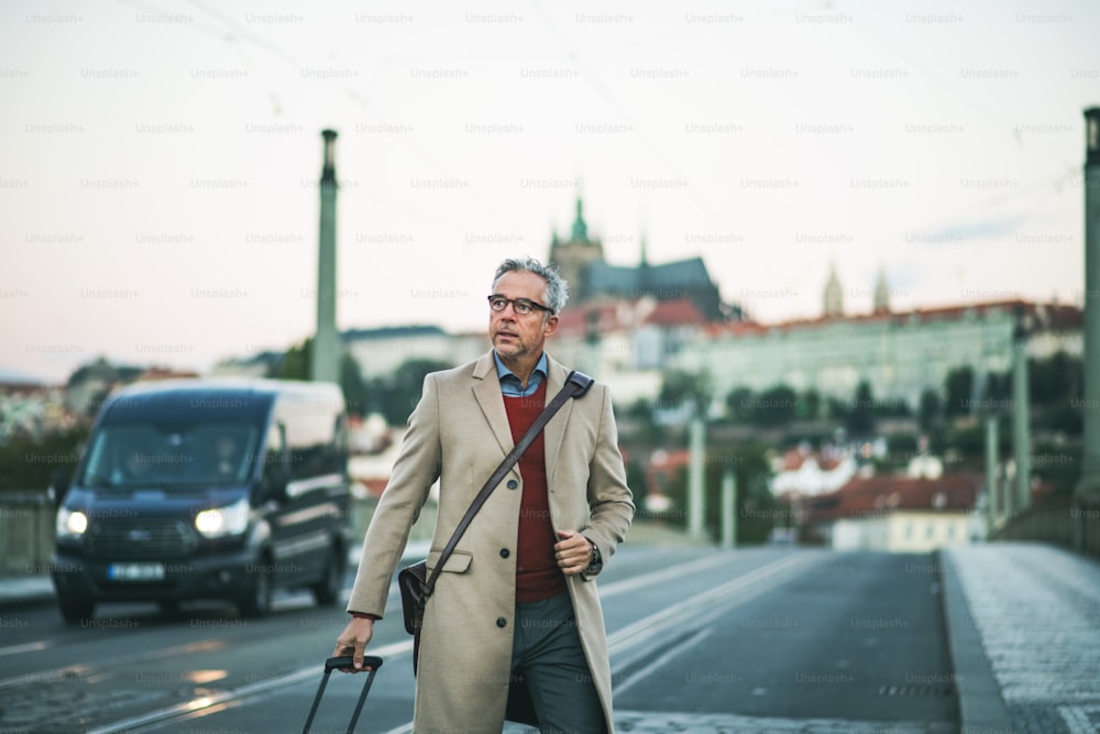 スーツケースを持つ成熟したハンサムな実業家がプラハ市の橋の上を歩き、道路を渡る。スペースをコピーします。