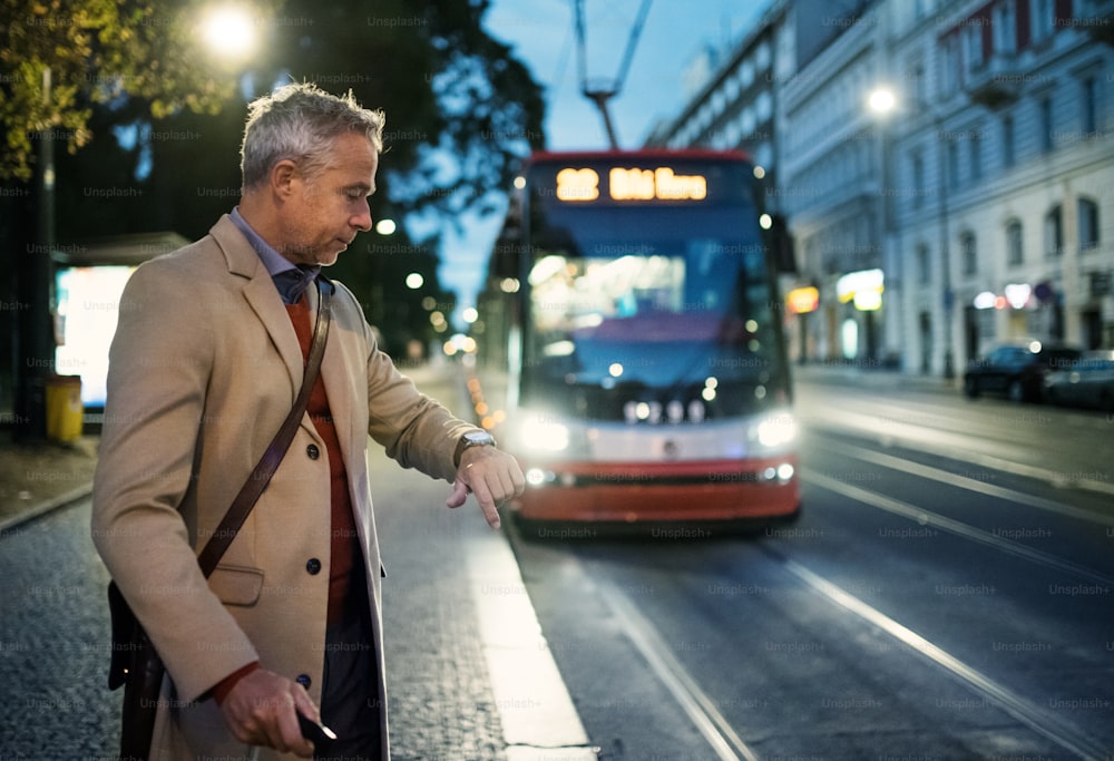 Mature businessman with suitcase waiting for a tram or a trolley car in the evening in Prague city, checking the time.