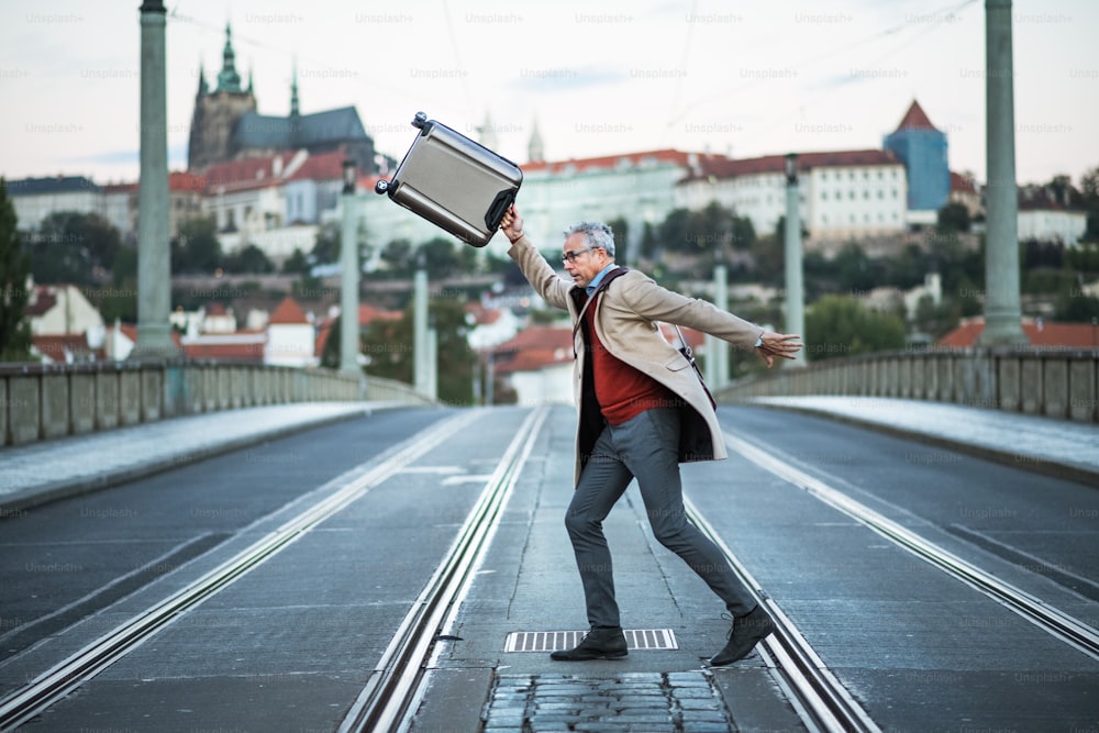 プラハ市の道路を横断するスーツケースを持つ幸せな成熟したハンサムなビジネスマン。