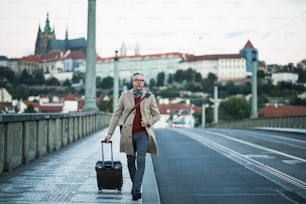 Mature handsome businessman with suitcase walking on a bridge in Prague city. Copy space.
