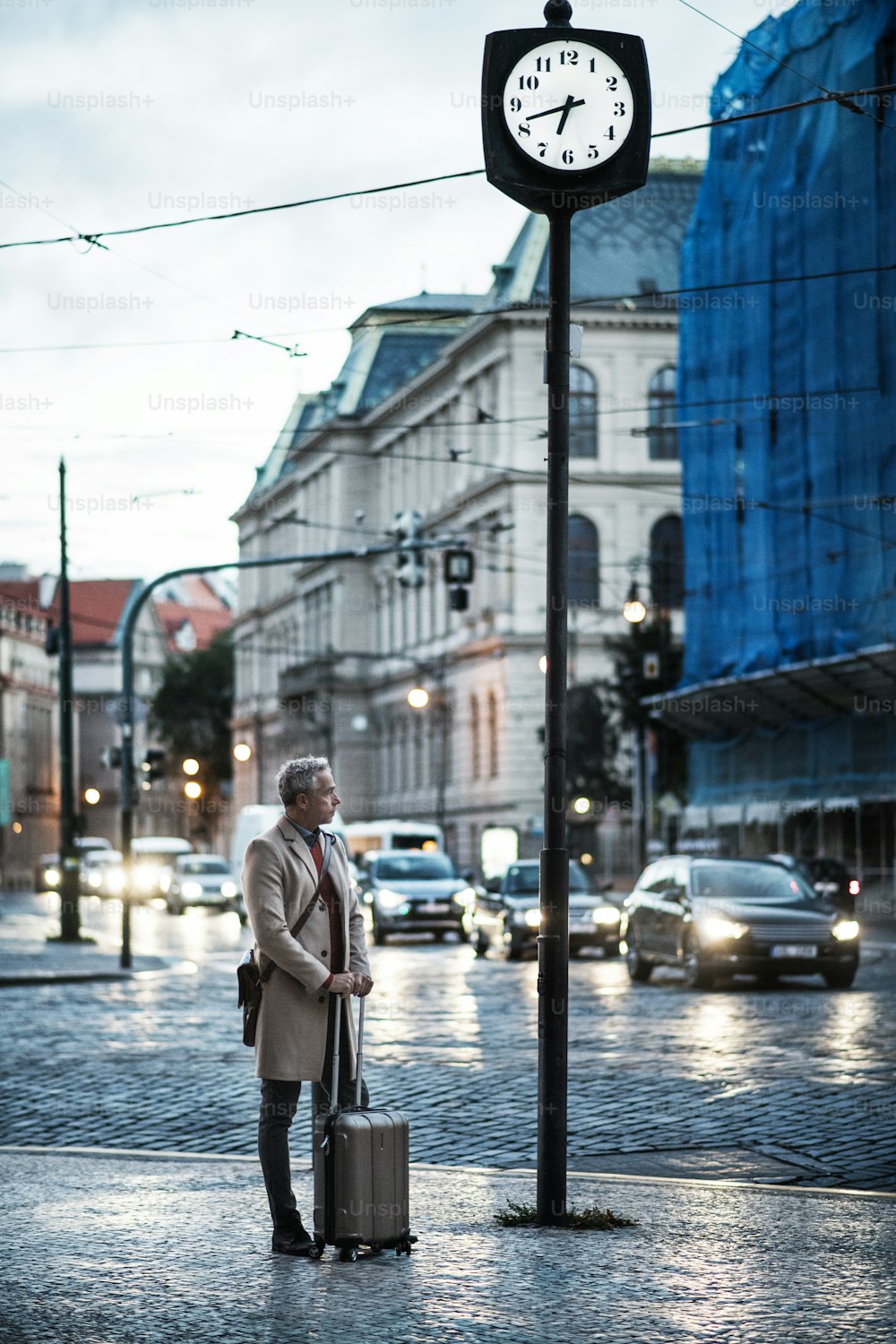 Mature handsome businessman with suitcase standing by the road early in the morning or evening.
