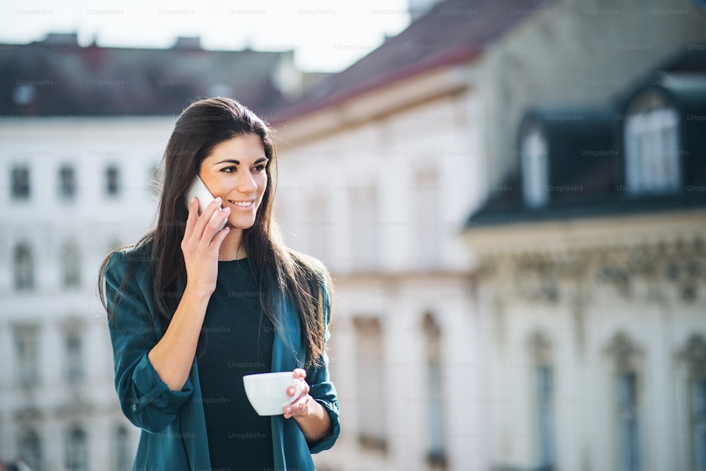 Young businesswoman with coffee and smartphone standing on a terrace outside an office in city, making a phone call. Copy space.
