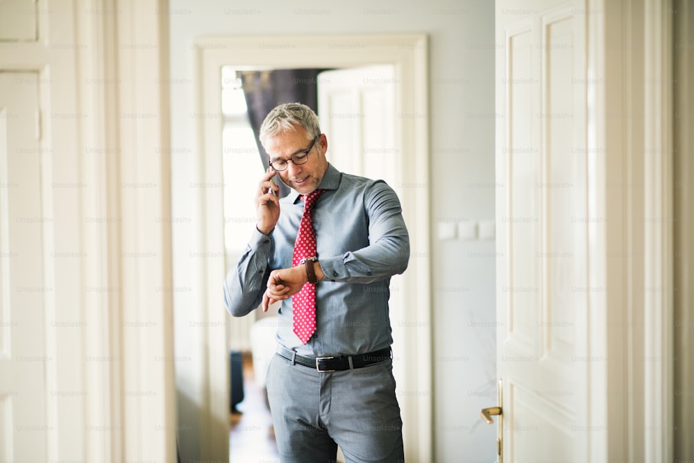 Mature businessman with glasses on a business trip standing in a hotel room, making phone call.