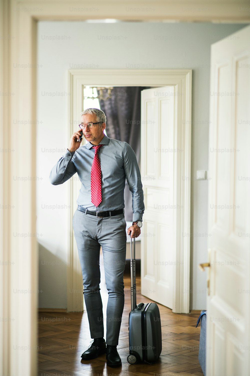Uomo d'affari maturo con occhiali e valigia in viaggio d'affari in piedi in una stanza d'albergo, usando lo smartphone. Copia spazio.