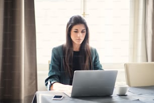 Young beautiful businesswoman sitting indoors at a table, using laptop.