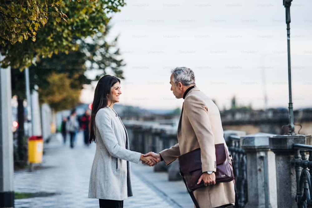 Un homme mûr et une jeune femme partenaires d’affaires debout à l’extérieur dans la ville de Prague, se serrant la main.