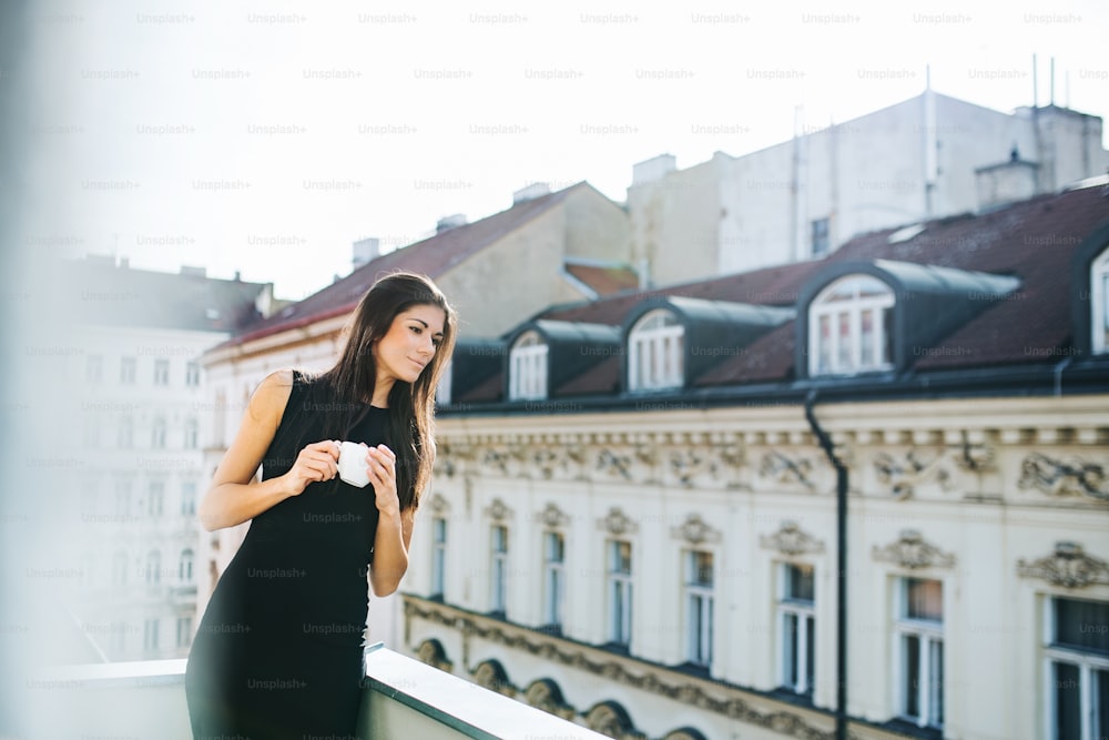 Giovane donna d'affari felice con una tazza di caffè in piedi su una terrazza fuori da un ufficio in città. Copia spazio.