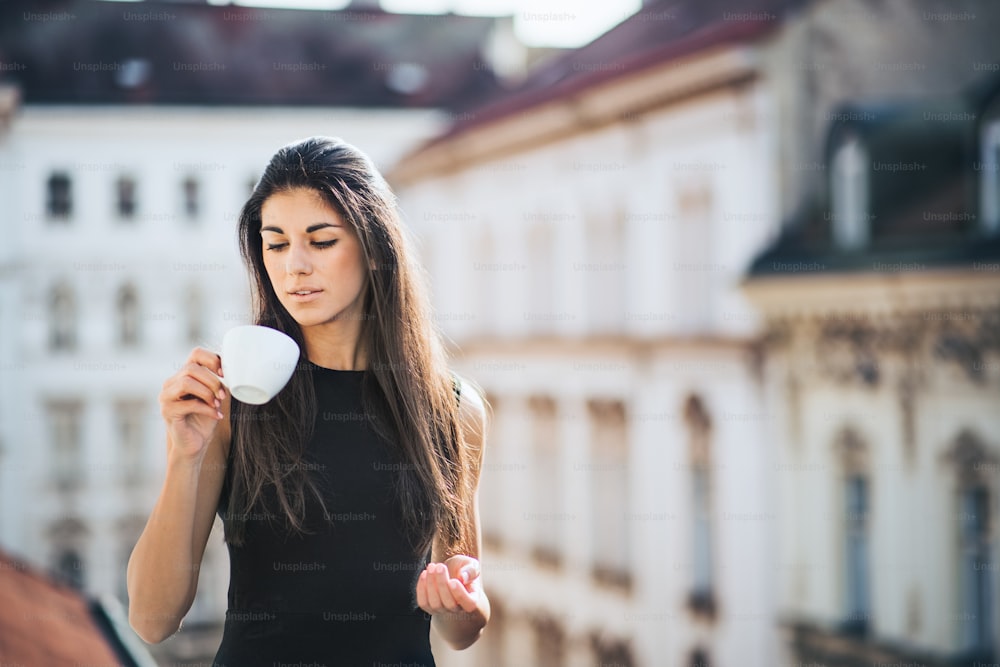 Giovane donna d'affari felice con una tazza di caffè in piedi su una terrazza fuori da un ufficio in città. Copia spazio.