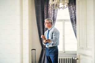 Mature businessman with glasses on a business trip standing in a hotel room, looking out of a window when getting dressed.