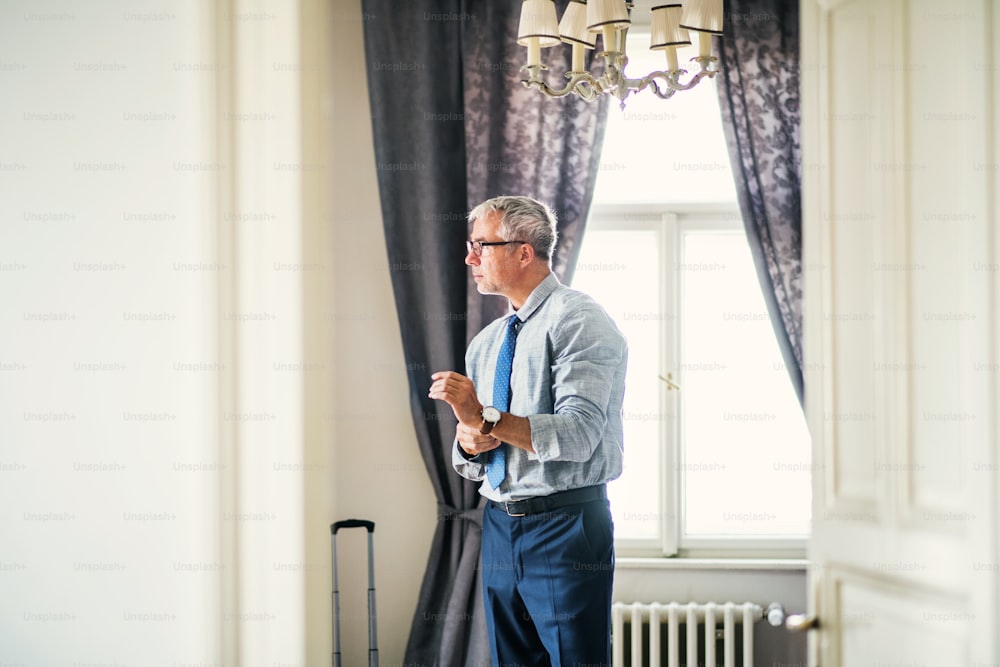 Empresário maduro com óculos em uma viagem de negócios em pé em um quarto de hotel, olhando por uma janela ao se vestir.