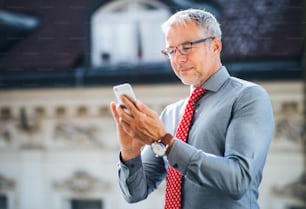 市内のオフィスのテラスに立ってテキストメッセージを送るスマートフォンを持つ成熟したビジネスマン。