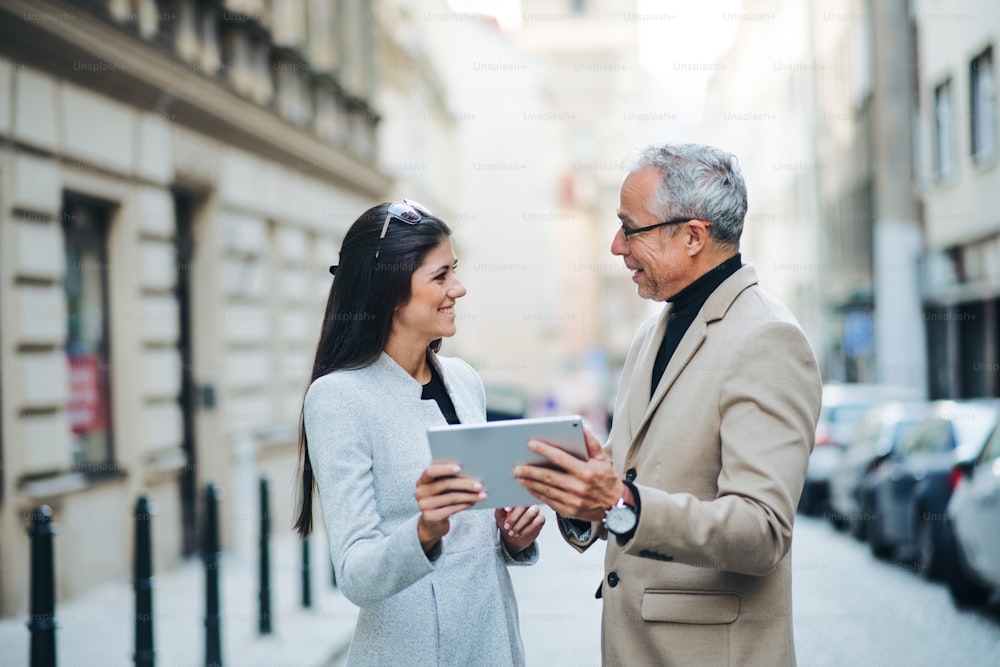 Un homme mûr et une jeune femme partenaires d’affaires avec une tablette debout à l’extérieur dans la ville de Prague, en train de parler.