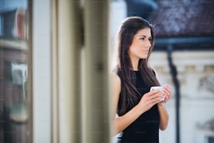 Happy young businesswoman with cup of coffee standing on a terrace outside an office in city. Copy space.