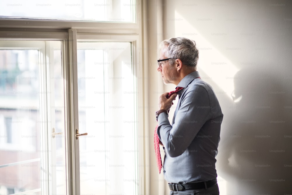 Mature businessman with glasses on a business trip standing in a hotel room, looking out of a window when getting dressed.