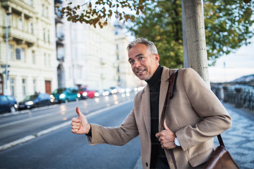 Mature businessman with bag standing on the street in city of Prague, raising his hand to hail a taxi cab.