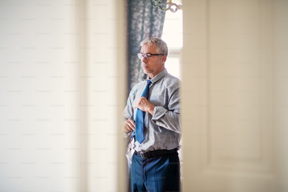 Mature businessman with glasses on a business trip standing in a hotel room, getting dressed.
