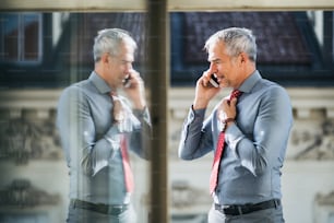 A mature businessman with smartphone standing on a terrace in an office in city, making a phone call.