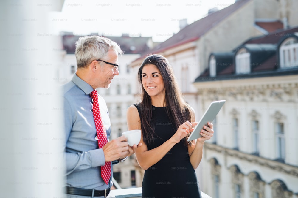 Hommes et femmes partenaires d’affaires avec tablette debout sur une terrasse dans un bureau en ville, en train de parler.