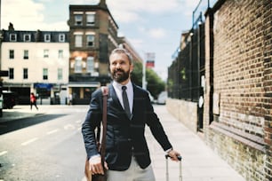 ロンドンの通りを歩くラップトップバッグとスーツケースを持つ流行に敏感なビジネスマン。スペースをコピーします。