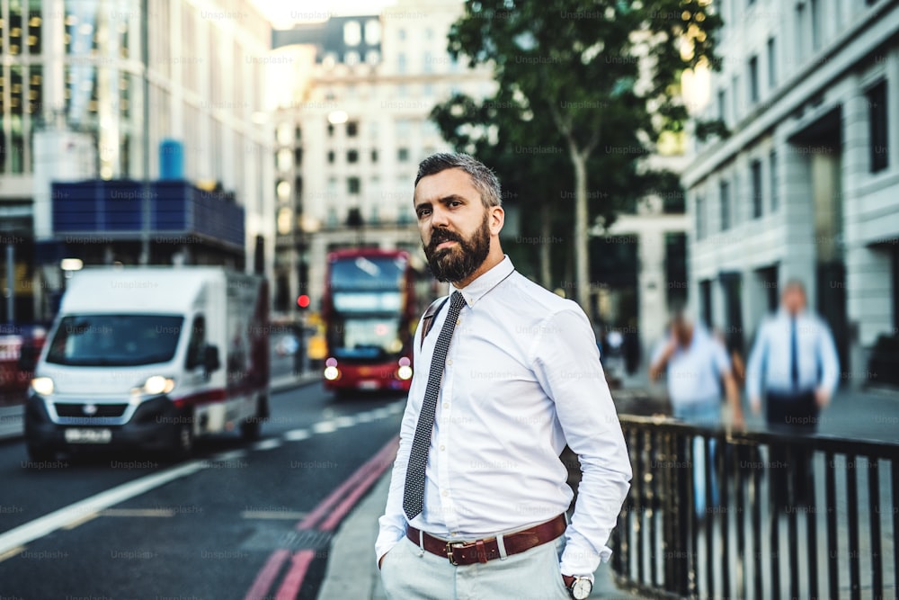 Hombre de negocios hipster parado en la calle en Londres junto a una calle concurrida, con las manos en los bolsillos.