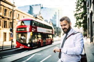 Hipster-Geschäftsmann, der in London auf der Straße steht und die Uhrzeit überprüft. Ein roter Doppeldeckerbus im Hintergrund.