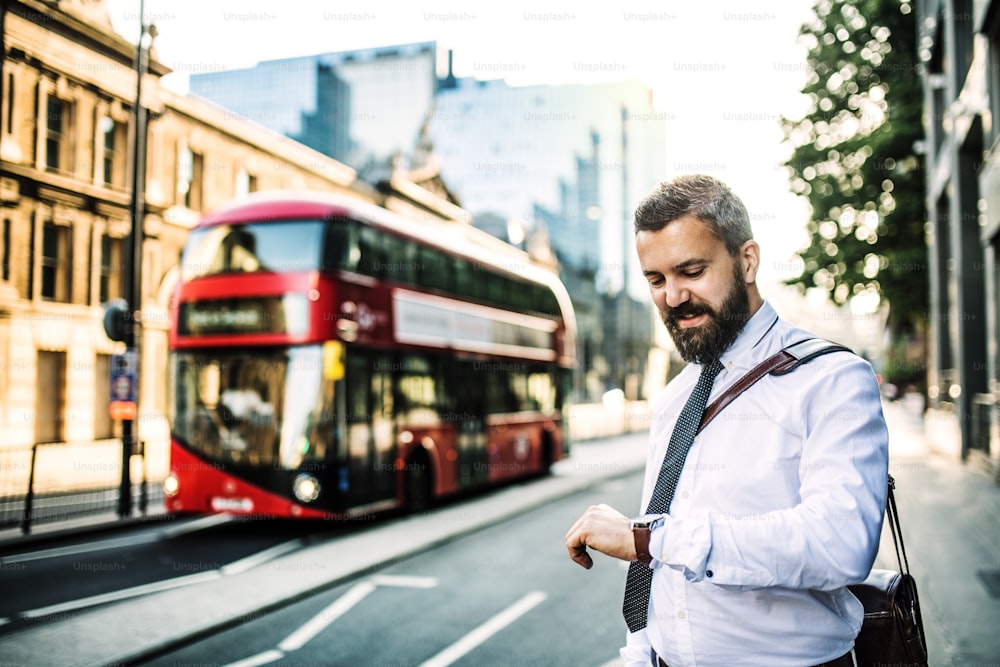 Hombre de negocios hipster parado en la calle en Londres, comprobando la hora. Un autobús de pisos douuble rojo en el fondo.