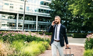 Uomo d'affari hipster con borsa per laptop, valigia e smartphone che cammina nel parco di Londra, facendo una telefonata.