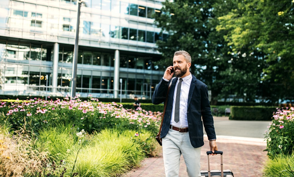 Hombre de negocios hipster con bolsa para computadora portátil, maleta y teléfono inteligente caminando en el parque de Londres, haciendo una llamada telefónica.