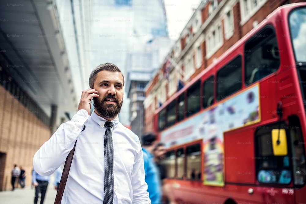 Hombre de negocios hipster con un teléfono inteligente caminando por la calle en Londres, haciendo una llamada telefónica.