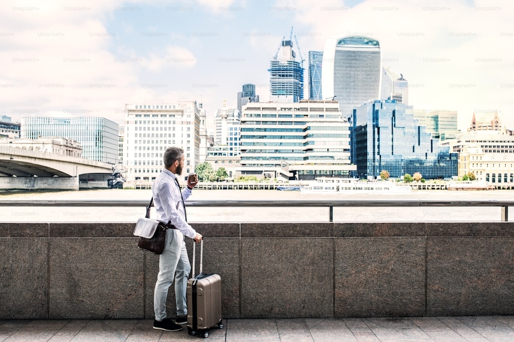 Homme d’affaires hipster avec valise debout au bord de la Tamise à Londres, tenant une tasse de café. Espace de copie.