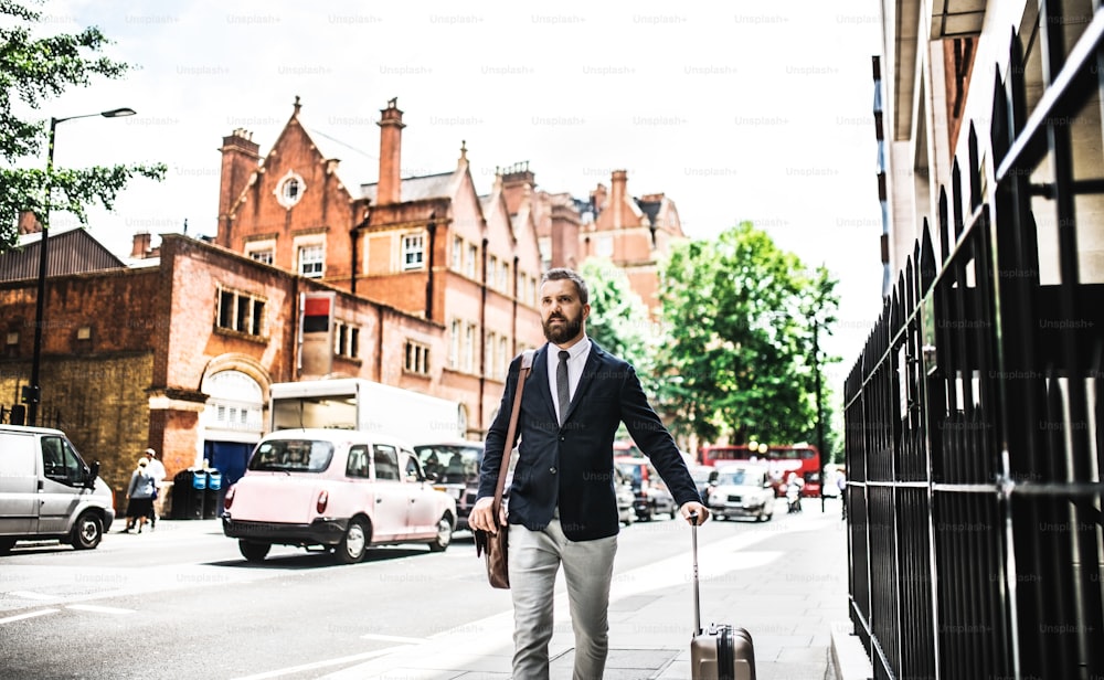 Homme d’affaires hipster avec un sac d’ordinateur portable et une valise marchant dans la rue à Londres. Espace de copie.