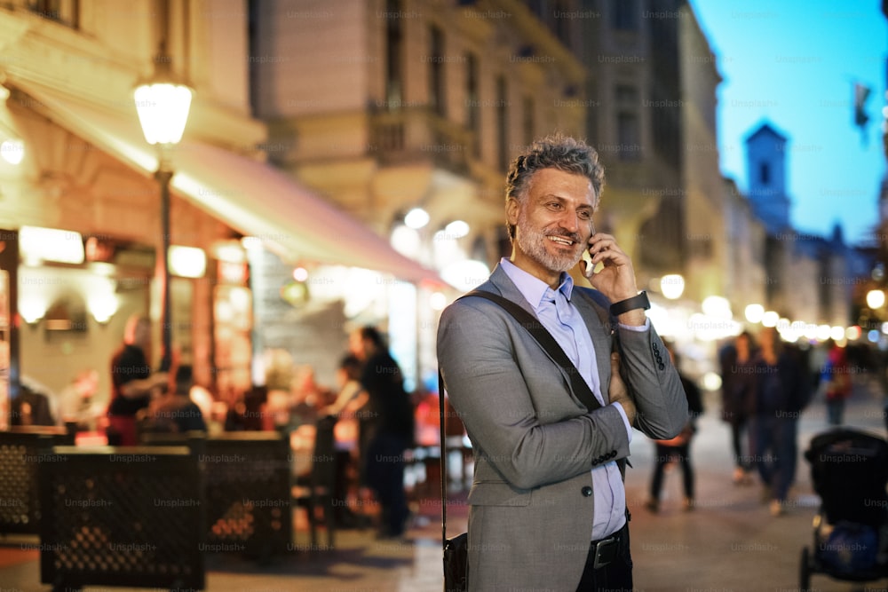 Bello uomo d'affari maturo con uno smartphone in una città la sera, facendo una telefonata.