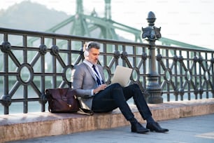 Handsome mature businessman with laptop in a city. Man sitting on a bridge, using laptop and headphones.
