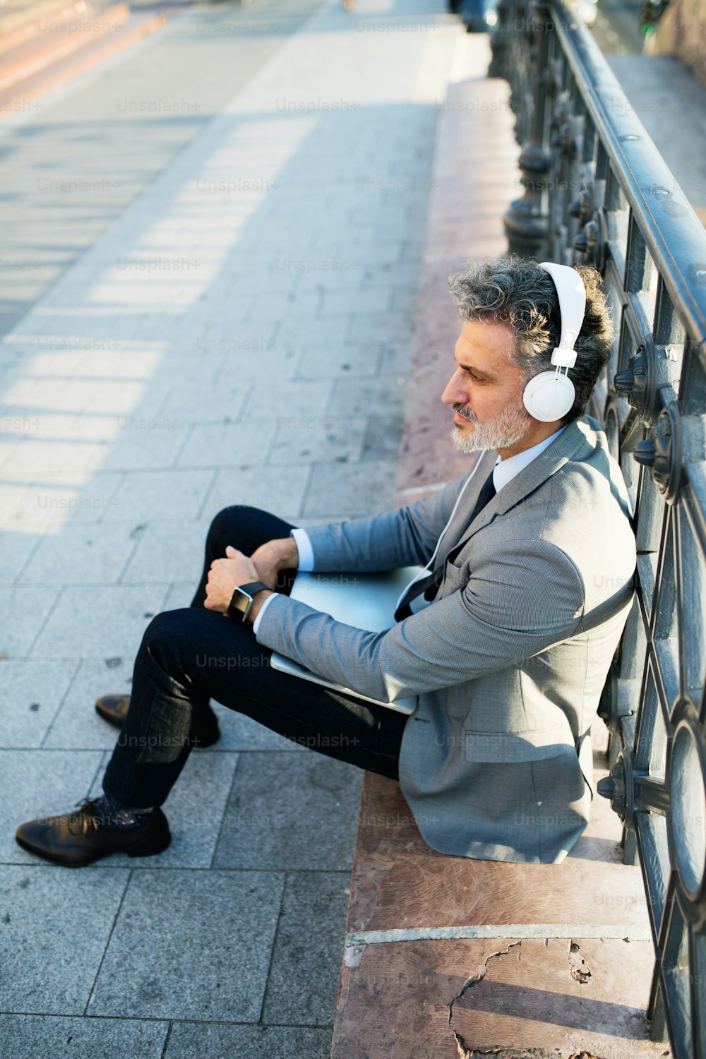 Guapo hombre de negocios maduro con computadora portátil en una ciudad. Hombre sentado en un puente, usando auriculares.