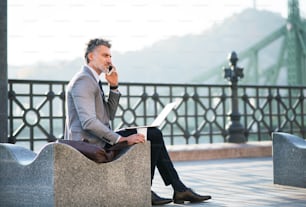 Handsome mature businessman with laptop and smartphone in a city. Man making a phone call.