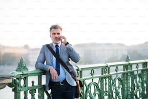 Handsome mature businessman with a smartphone in a city. Man standing on a bridge, making a phone call.
