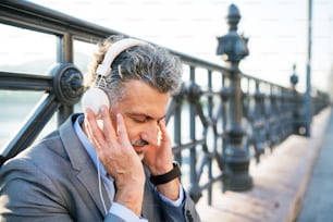 Bello uomo d'affari maturo con le cuffie in una città. Uomo seduto su un ponte, ascoltando musica.