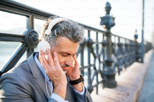 Handsome mature businessman with headphones in a city. Man sitting on a bridge, listening to music.